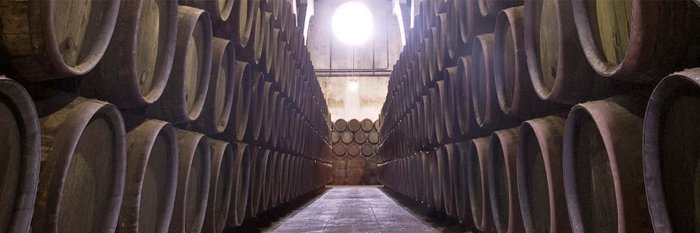 Ein langer Gang mit Holzfässern, die ordentlich aufgereiht sind, beleuchtet von einem runden Fenster am Ende.