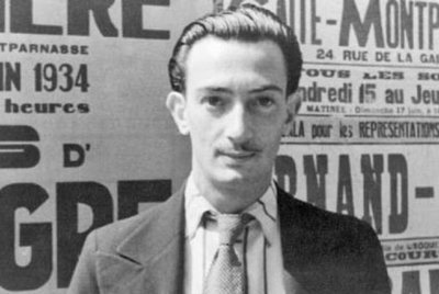 Ein junger Mann mit einem schmalen Gesicht und einem Schnurrbart steht vor einer Wand voller Plakate aus den 1930er Jahren.