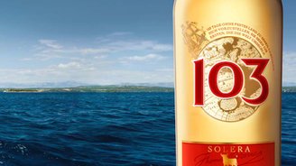 Ein Segelboot mit weißen Segeln gleitet über das blaue Wasser, während eine Flasche Osborne 103 im Vordergrund steht.