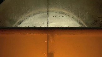 Durch ein großes, rundes Fenster ist eine leuchtend orangefarbene Flüssigkeit sichtbar, umgeben von grauen Wänden.