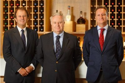 Drei Männer in Anzügen stehen vor einer Wand mit Weinregalen, die mit Flaschen gefüllt sind, und wirken selbstbewusst.