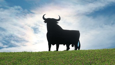Ein imposanter Stier steht auf einem sanften Hügel, umgeben von einer blühenden Wiese und einem strahlend blauen Himmel.