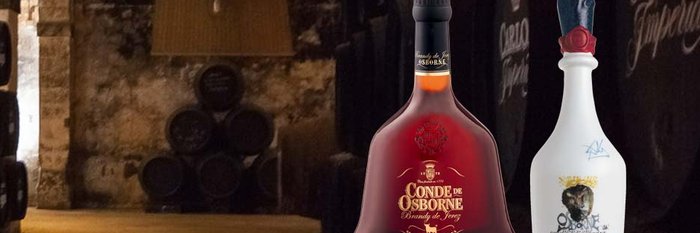 In einem dunklen Keller stehen große Fässer, während zwei Flaschen Brandy prominent im Vordergrund leuchten.