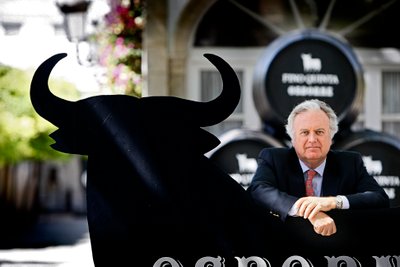Ein Mann in einem Anzug lehnt an einem großen, schwarzen Stier-Symbol vor einem Weingut mit Fässern im Hintergrund.