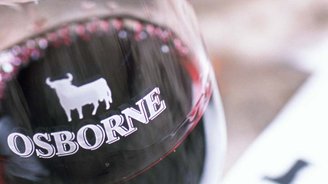 Nahaufnahme eines Weinglases mit 'OSBORNE'-Logo und Rind, umgeben von einem rustikalen Tisch und verschwommenen Papieren.